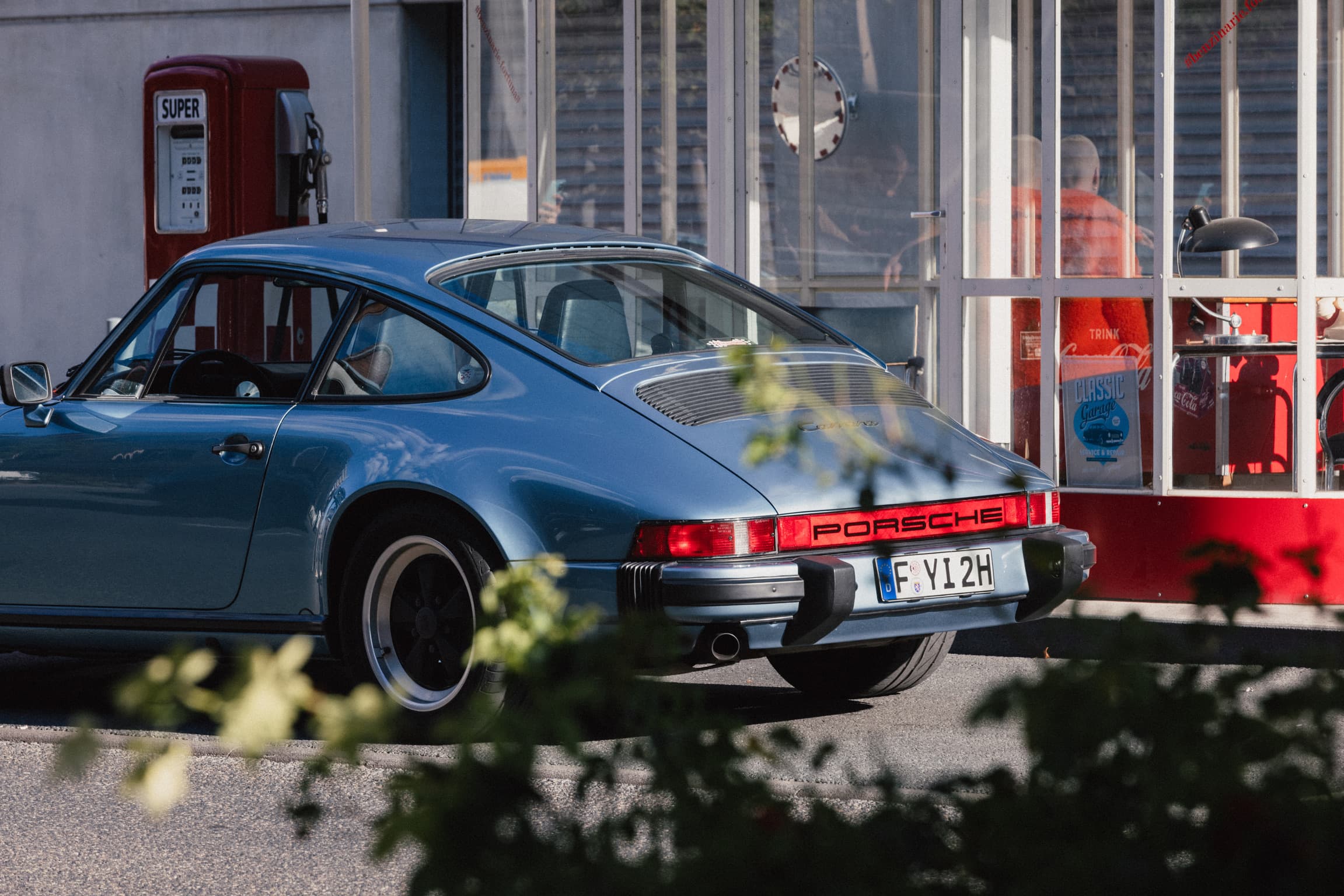 A blue classic Porsche 911 in front of an old petrol station
